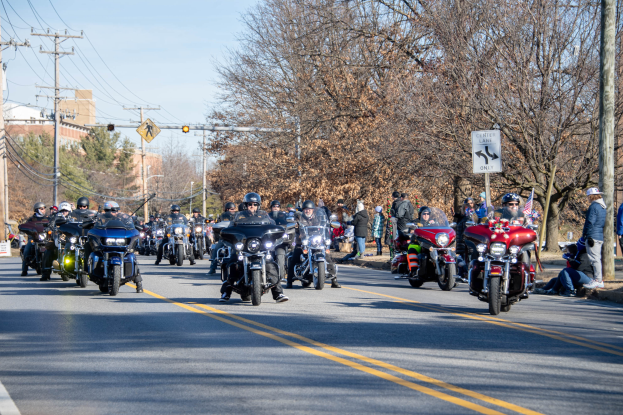 Eine Gruppe von Menschen auf Motorrädern, die eine Straße mit Strommasten, Schildern, Bäumen und Gebäuden entlangfahren, unter einem klaren blauen Himmel, wobei einige Fahrer Helme tragen.