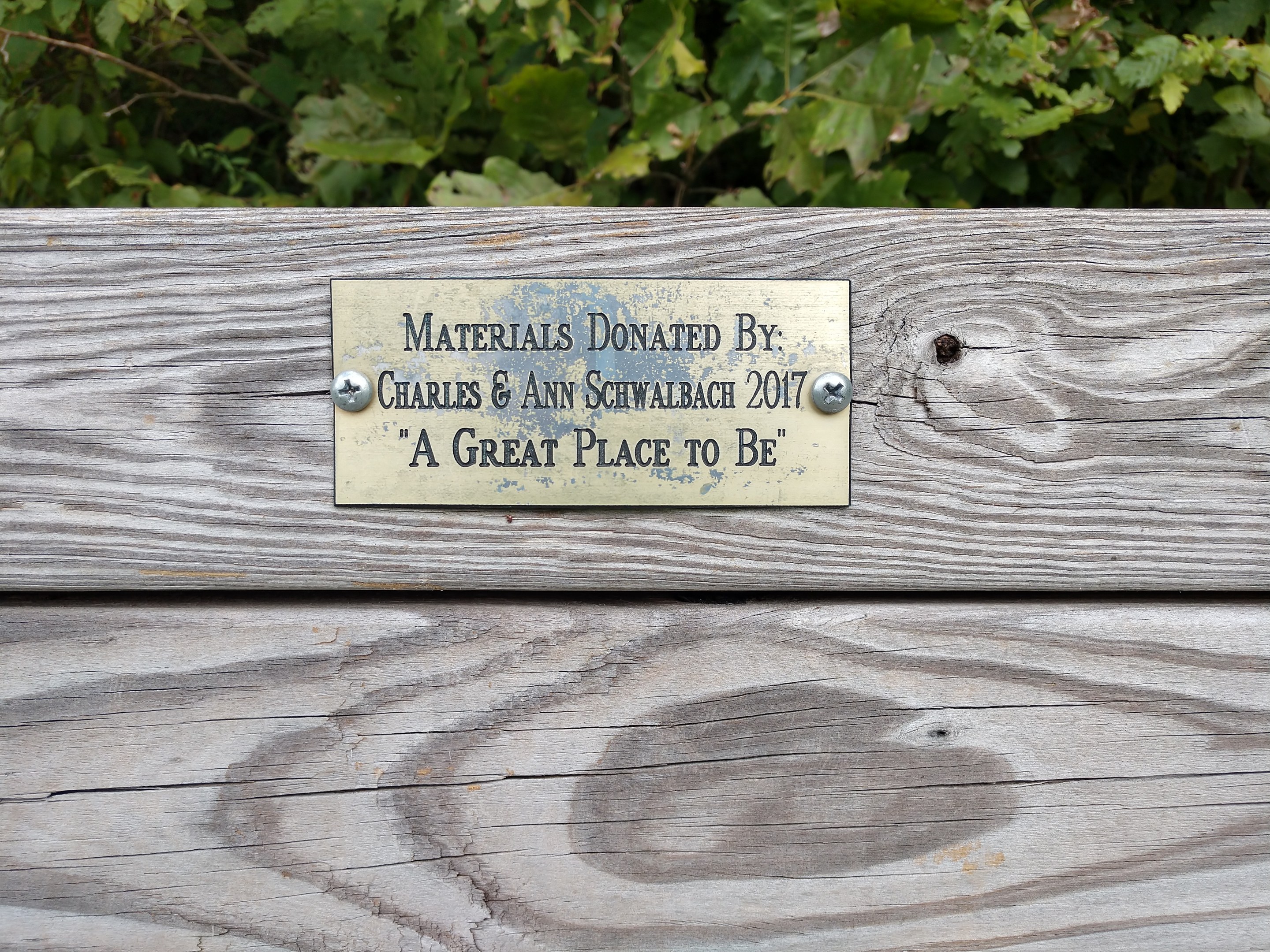 Holzbank mit einer Plakette, auf der "Materialien gespendet von Charles & Ann Schwabach 2017: Ein schöner Ort zu sein" steht, vor üppigen grünen Pflanzen.