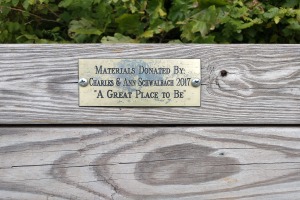 Holzbank mit einer Plakette, auf der "Materialien gespendet von Charles & Ann Schwabach 2017: Ein schöner Ort zu sein" steht, vor üppigen grünen Pflanzen.