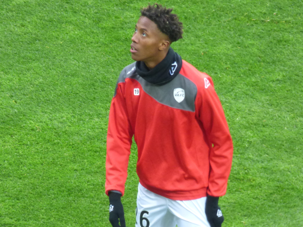 Ein junger Mann in roter Jacke und weißen Shorts steht auf einem Fußballfeld und trägt Baseballhandschuhe, sieht entschlossen nach rechts.