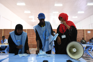 Eine Gruppe von Menschen um einen Tisch mit einem Megafon, Papieren und anderen Gegenständen versammelt, die scheinbar eine Wahllokal sind, mit anderen im Hintergrund unter Deckenlampen sitzend und stehend.