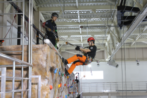 Zwei Personen in schwarzen und orangen Outfits und Helmen klettern an einer Felswand in einer Halle, gesichert durch Seile, mit einem Metallgeländer links und einer weißen Wand mit Leuchten im Hintergrund.