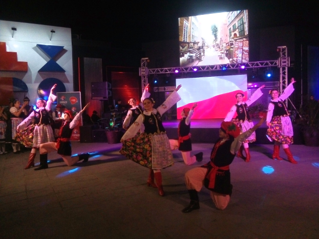 Eine Gruppe von Menschen tanzt auf einer Bühne vor einem großen Bildschirm, umgeben von einem sitzenden und stehenden Publikum, mit Blumentöpfen, Lichtern und anderen Gegenständen im Hintergrund.