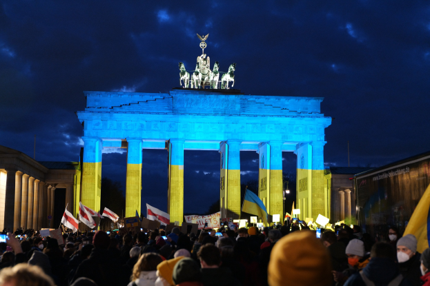 Menschenmenge mit Fahnen und Schildern vor dem Brandenburger Tor in Berlin, mit einer Banner auf der rechten Seite.