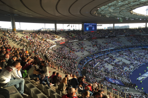Eine große Menschenmenge sitzt im Allianz Stadion in München, Deutschland, und schaut ein Fußballspiel. Rechts im Bild befindet sich eine Bühne mit ein paar Leuten darauf und im Hintergrund gibt es Fahnen, Stangen und einen Bildschirm. Der Himmel ist oben im Bild zu sehen und das Stadion scheint das Allianz Stadion in München, Deutschland, zu sein.