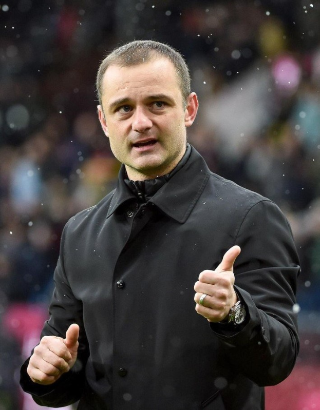 Fußballmanager in schwarzer Jacke mit Daumen-hoch-Geste, tragend eine Uhr, vor einem unscharfen Hintergrund.