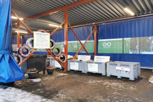 Großer Lagerraum mit verschiedenen Reifen, Containern, Topfpflanzen, bedruckten Platten, Pfählen, Beleuchtung und einem blauen Tuch, das eine Entsorgungsanlage mit Bäumen, Fahrzeugen und Schnee im Hintergrund anzeigt.