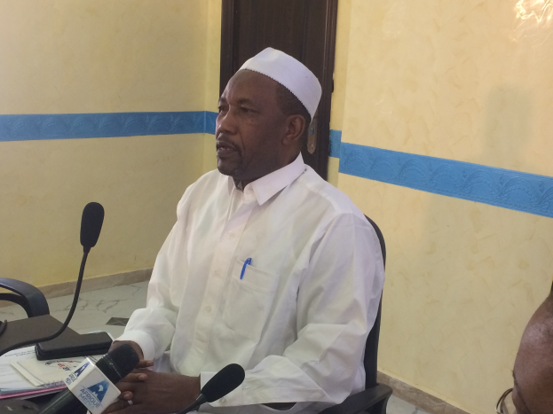 Ein Mann in einem weißen Kleid und einer Mütze sitzt an einem Tisch mit Mikrofonen, spricht, während Papiere und Gegenstände auf dem Tisch sichtbar sind, mit einer anderen Person mit Brille rechts von ihm, in einem Raum mit einer Wand und einer Tür im Hintergrund.