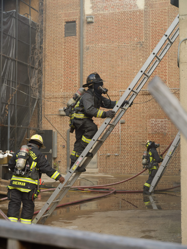 Feuerwehrleute in Helmen und Ausrüstung klettern eine Leiter vor einem Backsteingebäude mit Rohren auf dem Boden und einer Metallstange am Boden, mit einem anderen Gebäude und einem Netz im Hintergrund.