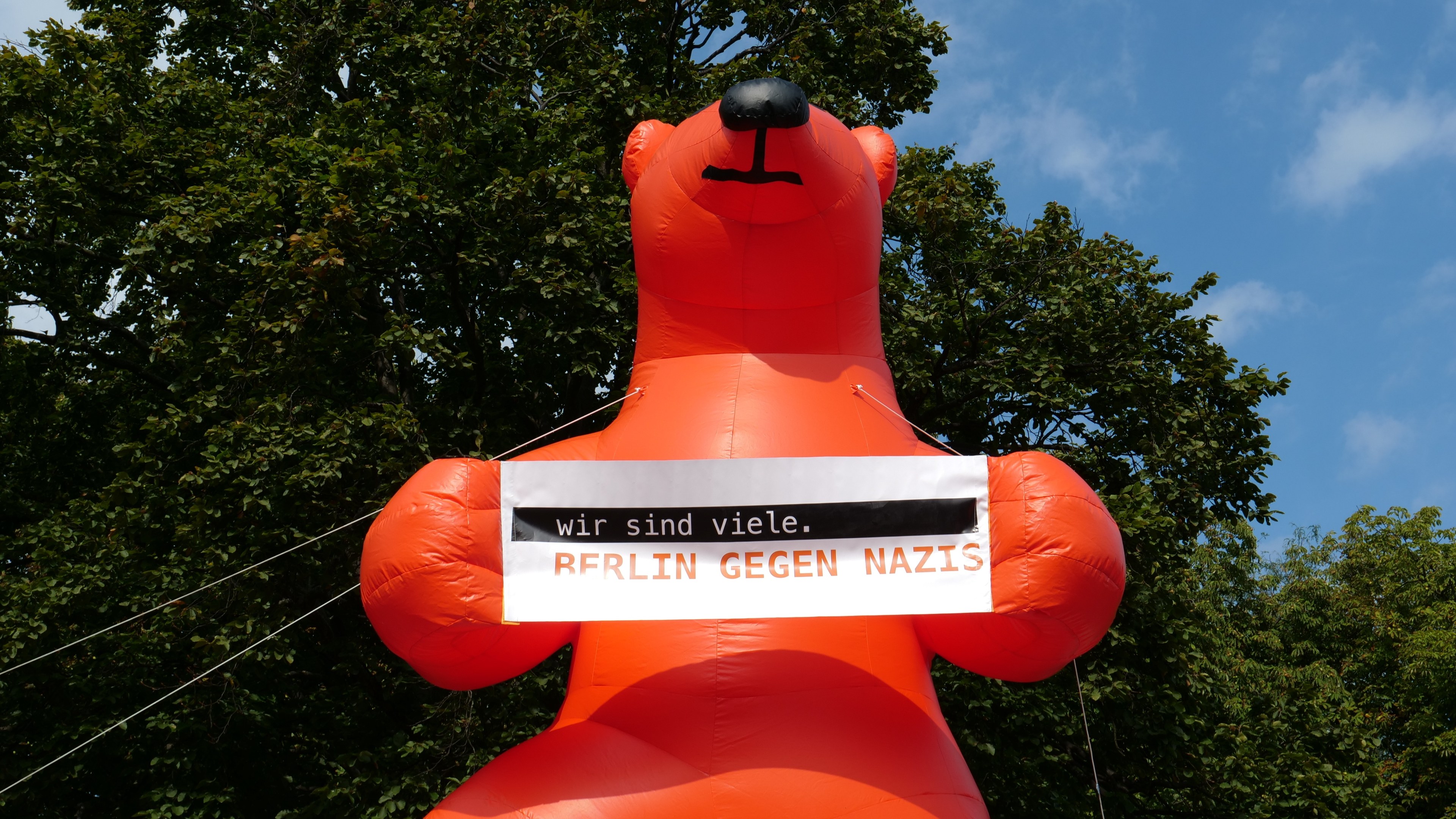 Aufblasbarer Bär mit einem Schild "Berlin gegen Nazis" vor einem Hintergrund aus Bäumen und Wolken.