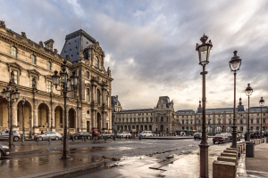 Außenansicht des Louvre-Museums in Paris mit seiner ikonischen Architektur, Straßenlaternen, Straßenlaternen, fahrenden Fahrzeugen, Fußgängern auf dem Bürgersteig und einer bewölkten Himmel.