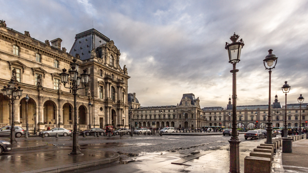 Außenansicht des Louvre-Museums in Paris mit seiner ikonischen Architektur, Straßenlaternen, Straßenlaternen, fahrenden Fahrzeugen, Fußgängern auf dem Bürgersteig und einer bewölkten Himmel.