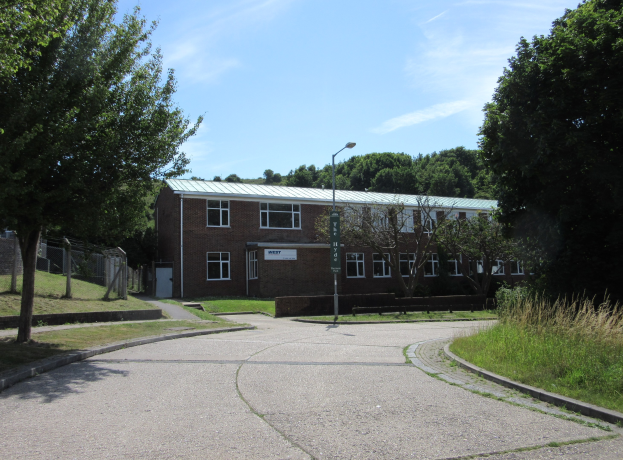Ein Schulgebäude mit Fenstern, einem Straßenschild, einem Schild, Bäumen, Gras, einem Zaun und einem bewölkten Himmel, das sich in einem Park mit saftigem Gras befindet.