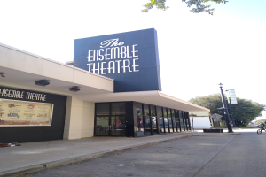 The Ensemble Theatre in San Francisco, ein modernes Gebäude mit Glaswänden und -türen, mit einer Schautafel, einer Person auf dem Gehweg, einem Fahrrad auf der Straße, einem Straßenschild, Bäumen und einem bewölkten Himmel.