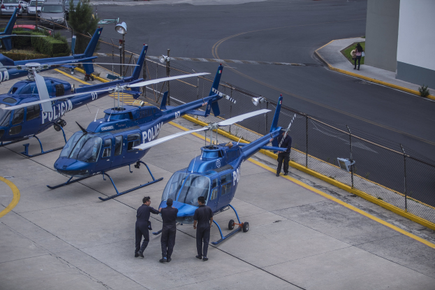 Gruppe von Polizeihubschraubern auf einem Flughafenvorfeld geparkt mit Menschen in der Nähe, ein Gebäude auf der rechten Seite und verschiedene Hintergrundelemente.