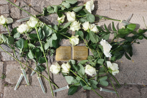 Weißer Rosenkranz mit Kerzen und goldenen Gegenständen mit Inschrift vor einem Berliner Denkmal.