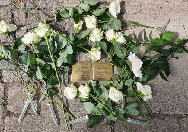 Weißer Rosenkranz mit Kerzen und goldenen Gegenständen mit Inschrift vor einem Berliner Denkmal.