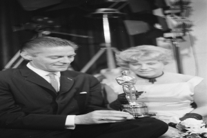 Ein Mann in einem schwarzen Blazer, einem weißen Hemd und einer Krawatte hölt eine Oscar-Statue, waehrend er neben einer Frau in Abendgarderobe sitzt, die einen Blumenstrauß hölt, mit Lichtern im Hintergrund, alles in Schwarz-Weiß.