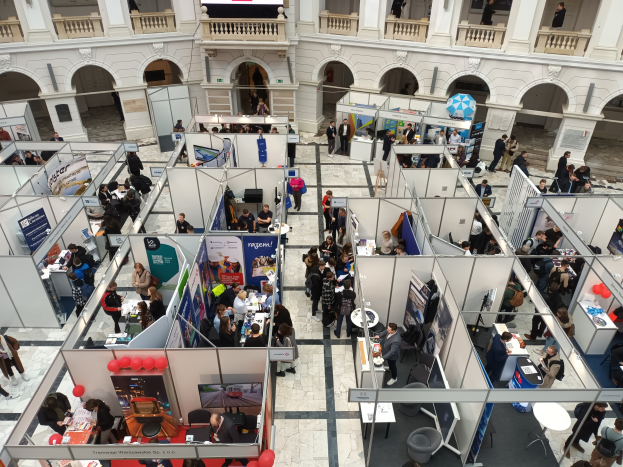 Eine große Gruppe von Menschen auf einer internationalen Fachmesse mit Ständen, Bannern, Tischen, Stühlen und Ballons in einem Raum mit gewölbter Architektur und einem Bildschirm im Hintergrund.