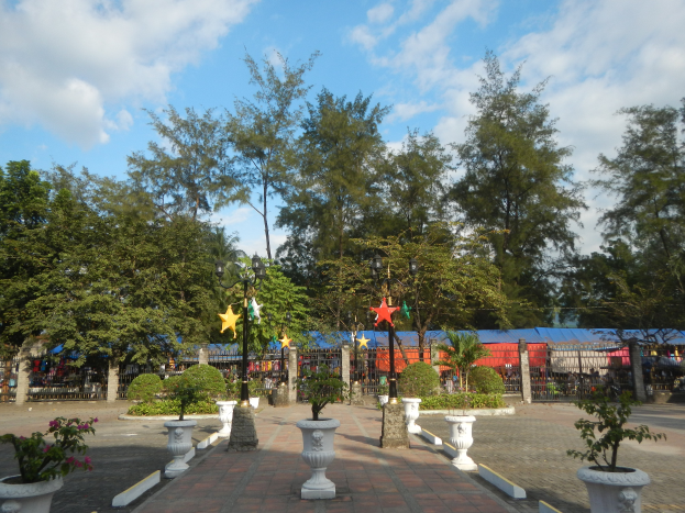 Ein Park mit Topfpflanzen und Bäumen in der Mitte, umgeben von Straßenlaternen, Schmuckelementen, Grills und einem bewölkten Himmel im Hintergrund.