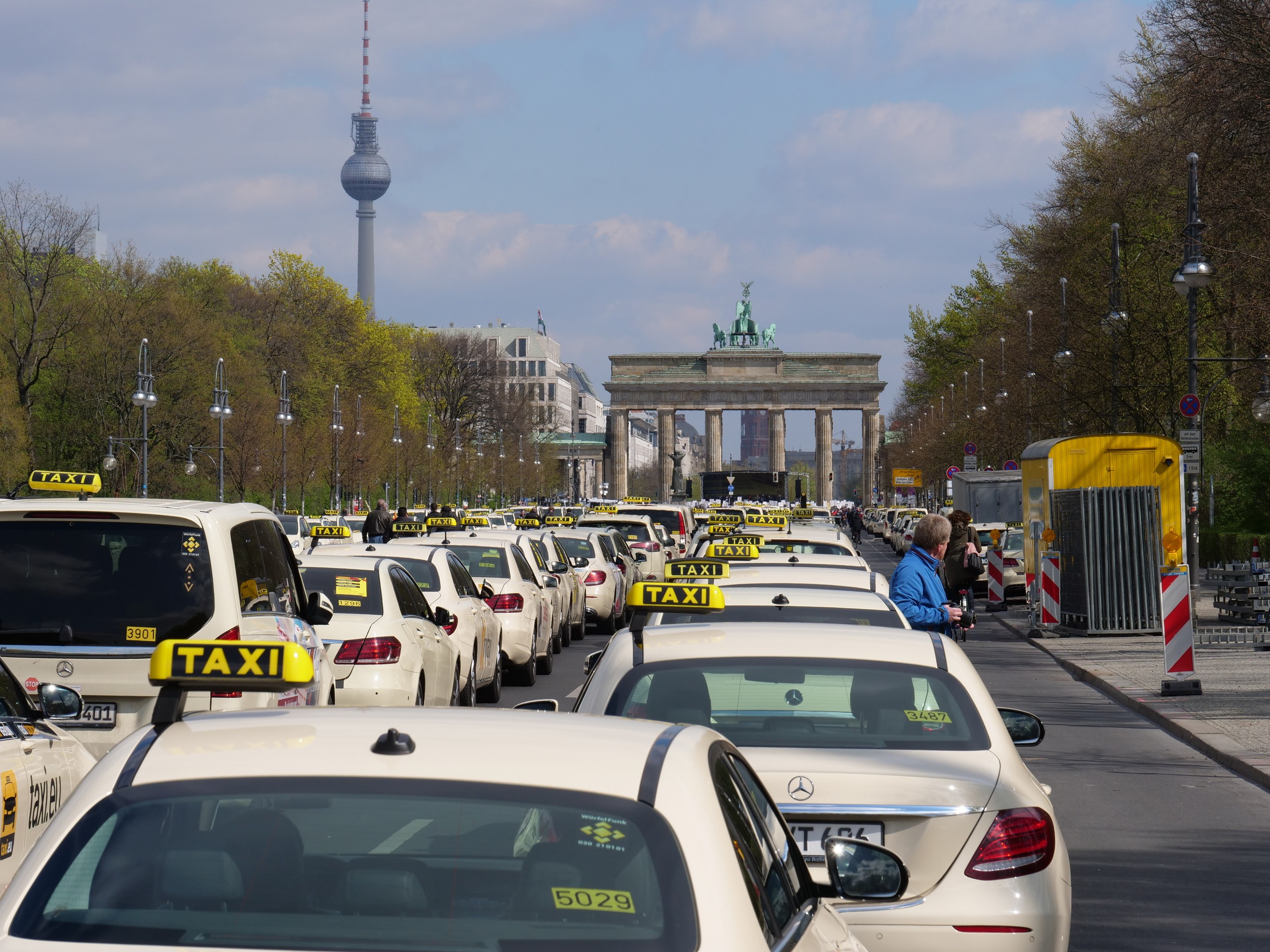 Eine belebte Straße in Berlin mit zahlreichen geparkten Taxis, Fußgängern auf dem Gehweg, Laternen, Bäumen, Gebäuden, einem fernen Bogen mit Statuen und einem Turm.