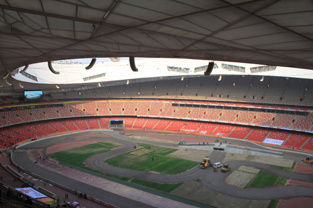 Großes Stadion voller Zuschauer, Fahrzeuge auf der Straße, grüner Rasen, Texttafeln, Geländer, Deckenbeleuchtung und ein Bildschirm.