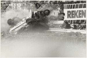 Ein Schwarz-Weiß-Foto eines demolierten Rennwagens, umgeben von Reifen, mit einer Gruppe von Menschen auf der rechten Seite und einer Tafel mit Text im Hintergrund.
