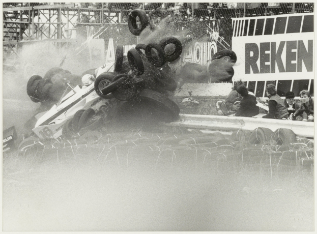 Ein Schwarz-Weiß-Foto eines demolierten Rennwagens, umgeben von Reifen, mit einer Gruppe von Menschen auf der rechten Seite und einer Tafel mit Text im Hintergrund.