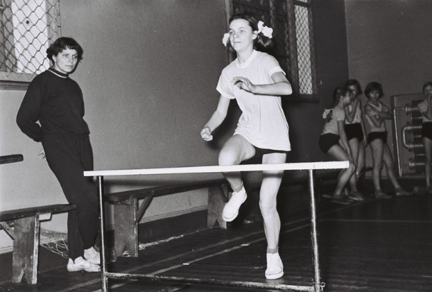 Schwarzes und weißes Foto einer Frau, die über ein Hindernis in einer Turnhalle springt, mit Zuschauern und Fenstern im Hintergrund.