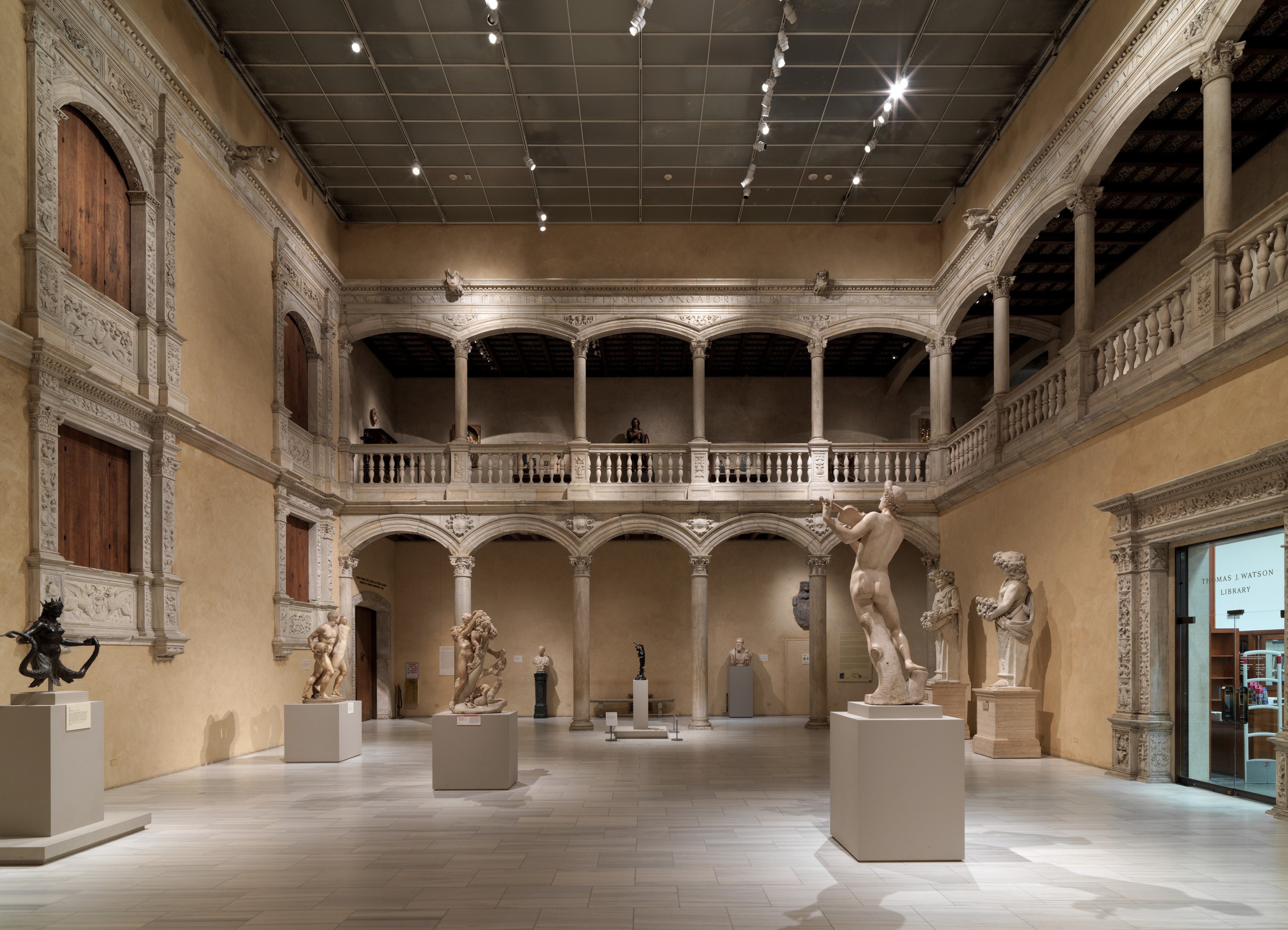Ein geräumiger Museumsgalerie mit zahlreichen Statuen und Skulpturen auf Sockeln, mit architektonischen Elementen wie Wänden, Säulen, Geländern und einer beleuchteten Decke und einigen Besuchern, die die Kunstwerke betrachten.