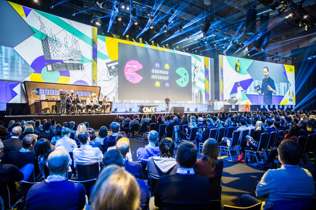 Eine große Menschenmenge, die auf Stühlen vor einer Bühne sitzt, mit einigen Individuen, die auf dem Podium in der Vordergrund stehen, einem Tisch mit verschiedenen Gegenständen links und Bildschirmen und Lichtern im Hintergrund auf der Deutschen Internet-Konferenz 2019.