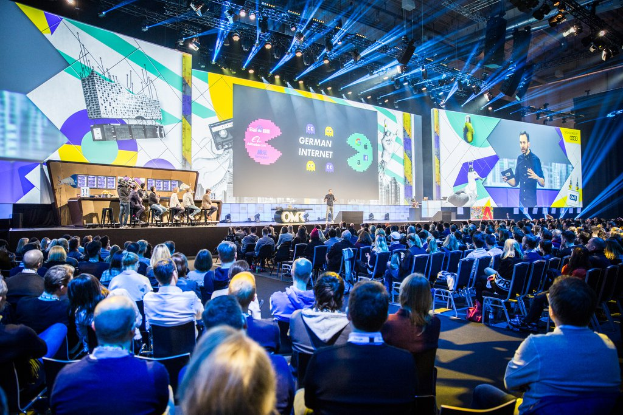 Eine große Menschenmenge, die auf Stühlen vor einer Bühne sitzt, mit einigen Individuen, die auf dem Podium in der Vordergrund stehen, einem Tisch mit verschiedenen Gegenständen links und Bildschirmen und Lichtern im Hintergrund auf der Deutschen Internet-Konferenz 2019.