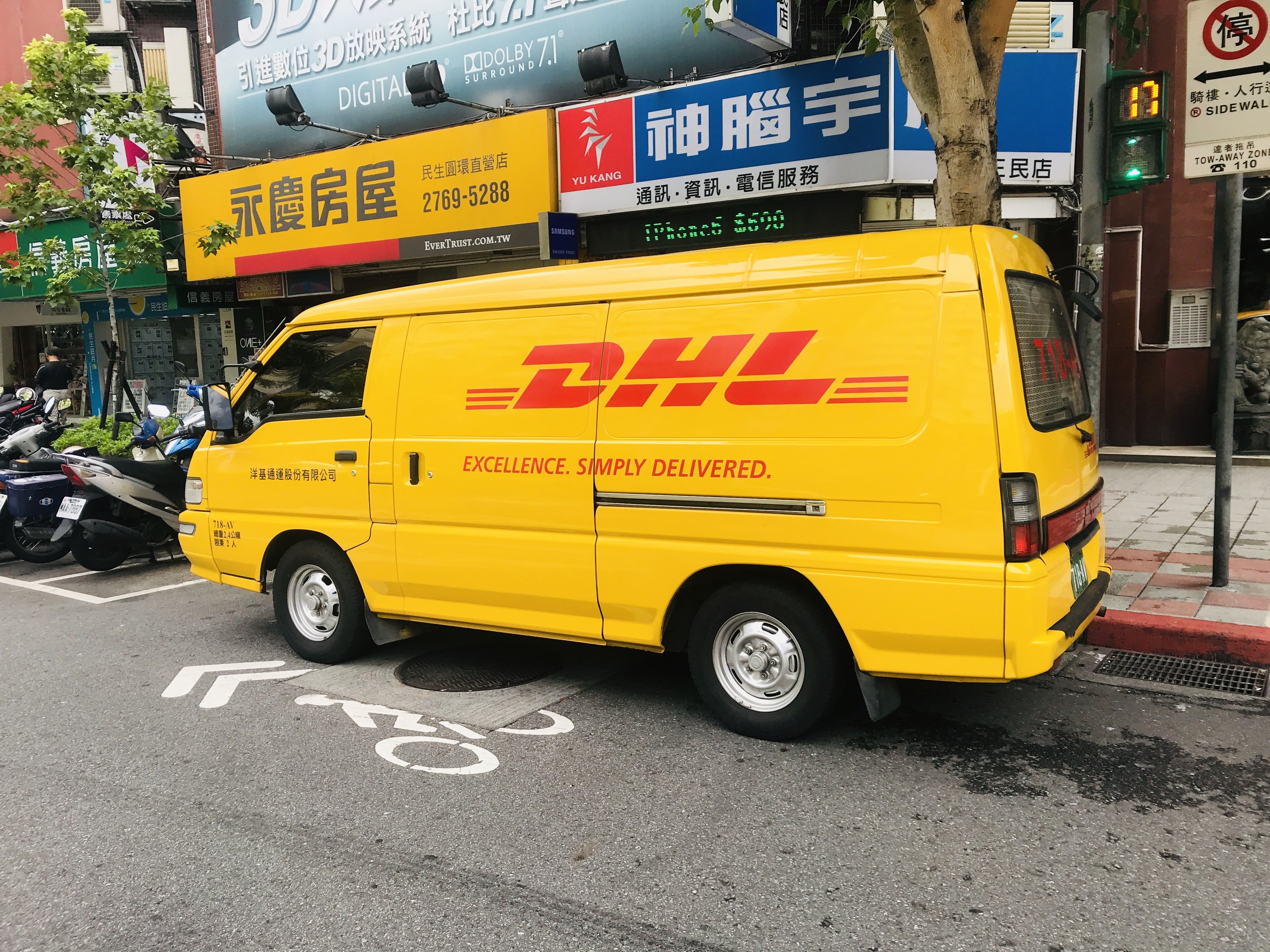 Gelbes DHL-Lieferfahrzeug auf der Straßenseite mit Gebäuden, Bäumen, Texttafeln, Masten, Verkehrszeichen und einer Person auf dem Gehweg.