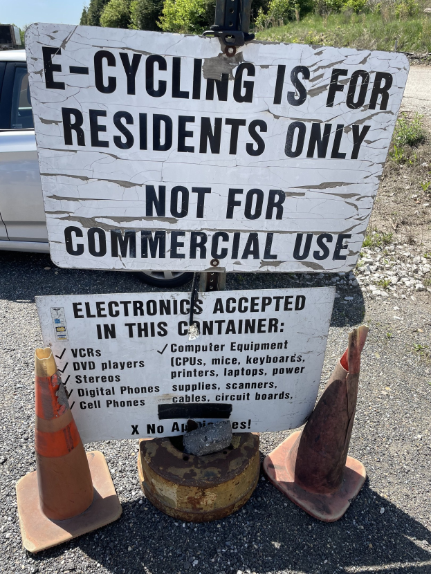 Schild mit der Aufschrift 'e-Bike-Fahren nur für Bewohner' neben einer Straße mit Absperrgittern, einem Fahrzeug, Bäumen und einem klaren blauen Himmel im Hintergrund.