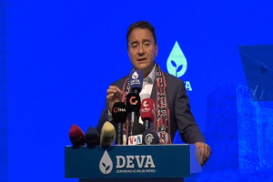 Ein Mann steht an einem Podium mit Mikrofonen, trägt einen Mantel und eine Schärpe und hält ein Schild mit der Aufschrift "deva-Geschäftsführer sagt, es sei keine gute Idee für die Zukunft", ein Objekt und ein Bildschirm im Hintergrund.