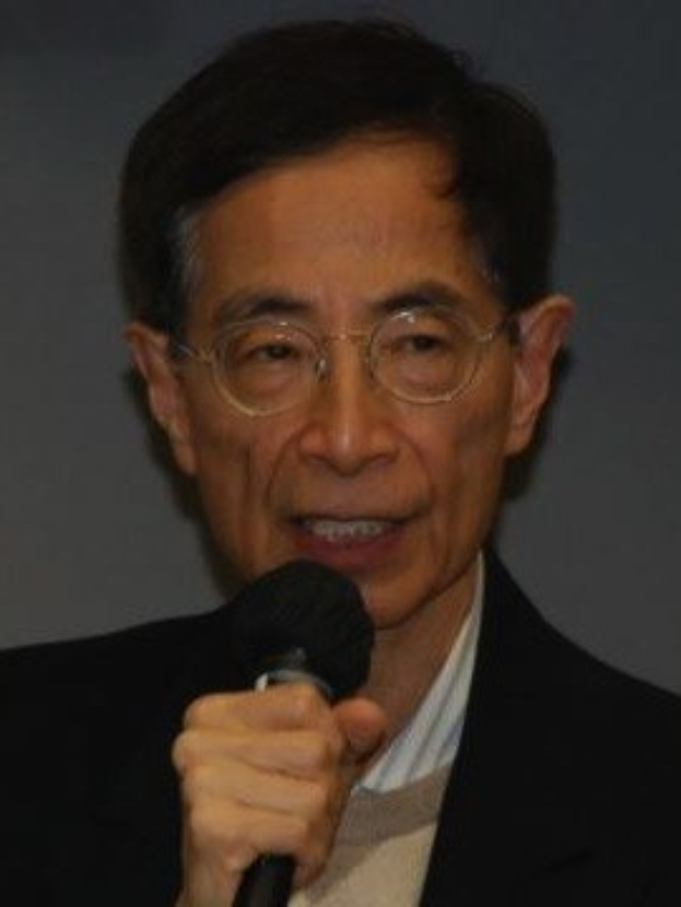 Ein Mann in einem schwarzen Blazer und Brille hält ein Mikrofon mit ernster Miene, was darauf schließen lässt, dass er Ökonom ist.