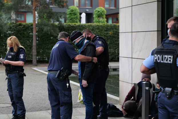 Deutsche Polizeibeamte stehen in einer Gruppe vor einem Gebäude mit einer Frau, die ein Buch und einen Stift hält, während Menschen in der Nähe sitzen und stehen, mit Bäumen und Gebäuden im Hintergrund.