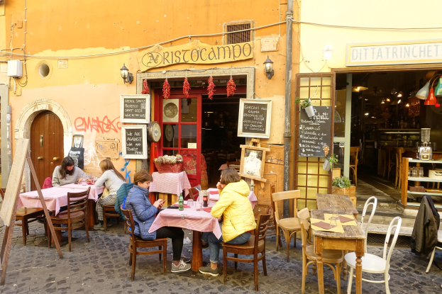 Eine Gruppe von Menschen, die an Tischen vor einem Restaurant in Rom, Italien, sitzen, mit Flaschen, Gläsern und anderen Gegenständen auf den Tischen, Wände mit Texttafeln geschmückt, eine Tür nach innen führend und beleuchtete Lampen, mit Regalen voller verschiedener Gegenstände im Inneren.