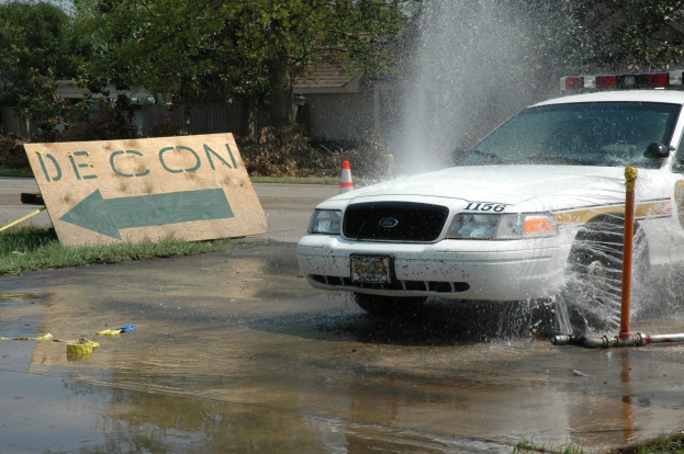 Ein Polizeiwagen fährt durch eine Pfütze, mit einem Schild, einer Verkehrskegel und anderen Gegenständen am Boden, mit Bäumen, Gebäuden und einem klaren blauen Himmel im Hintergrund.