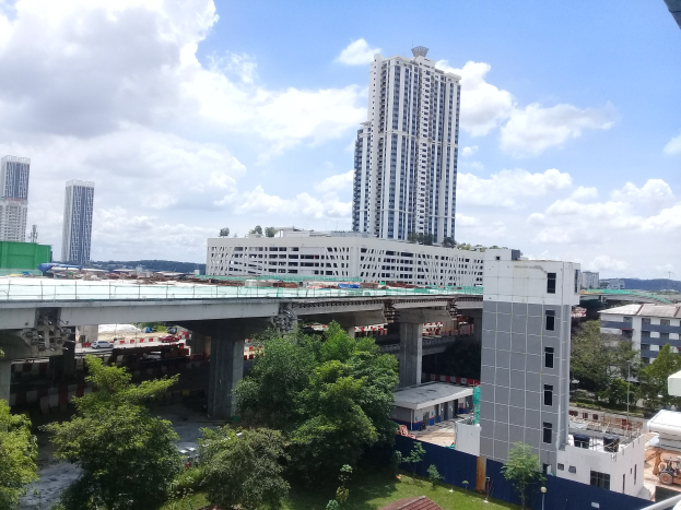Luftaufnahme einer Stadt mit Gebäuden, Bäumen, Gras, Fahrzeugen, einer Brücke, Masten und bewölktem Himmel, die eine Baustelle für eine neue Wohnanlage zeigt.