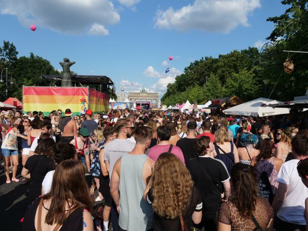 Eine große Menge Menschen geht eine Straße mit Zelten, Bäumen, Pfählen, Lichtern und einer Statue entlang während des Christopher Street Day Festivals in Berlin, mit Gebäuden und einem Himmel voller Wolken und Ballons im Hintergrund.