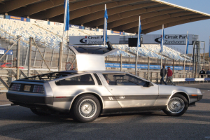 Ein silberner DeLorean DMC-12 fährt auf einer Straße umgeben von Menschen, Fahnen, Schildern, Geländern, Stühlen und einem Schuppen, mit Bäumen und einem klaren blauen Himmel im Hintergrund.