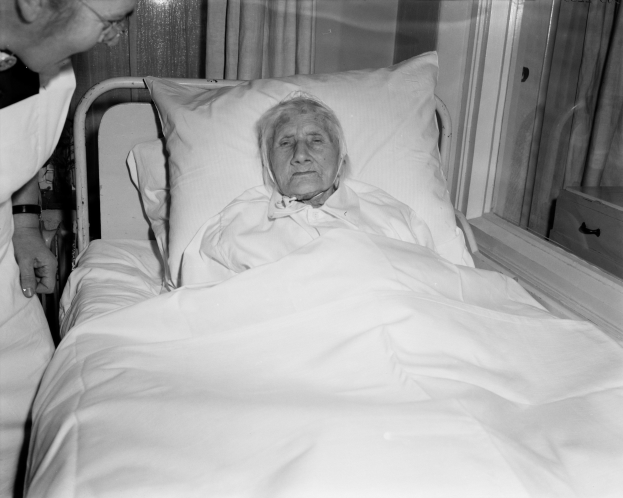 Ein älteres Frau liegt in einem Krankenhausbett mit einer weißen Bettdecke und Kissen, während eine Krankenschwester neben ihr steht, mit einem Fenster und Vorhängen im Hintergrund.