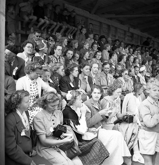 Schwarzes und weißes Foto einer Menge, die in einem Stadion mit einer hölzernen Wand und einem Dach im Hintergrund sitzt, einige halten Kameras.