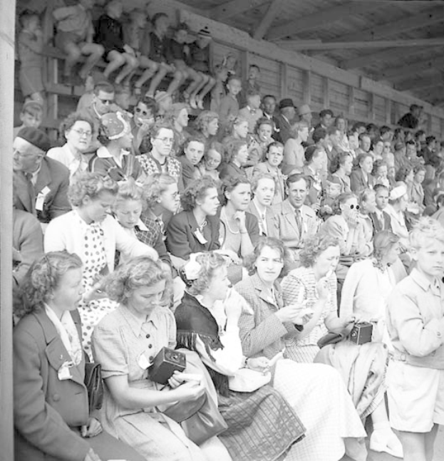 Schwarzes und weißes Foto einer Menge, die in einem Stadion mit einer hölzernen Wand und einem Dach im Hintergrund sitzt, einige halten Kameras.