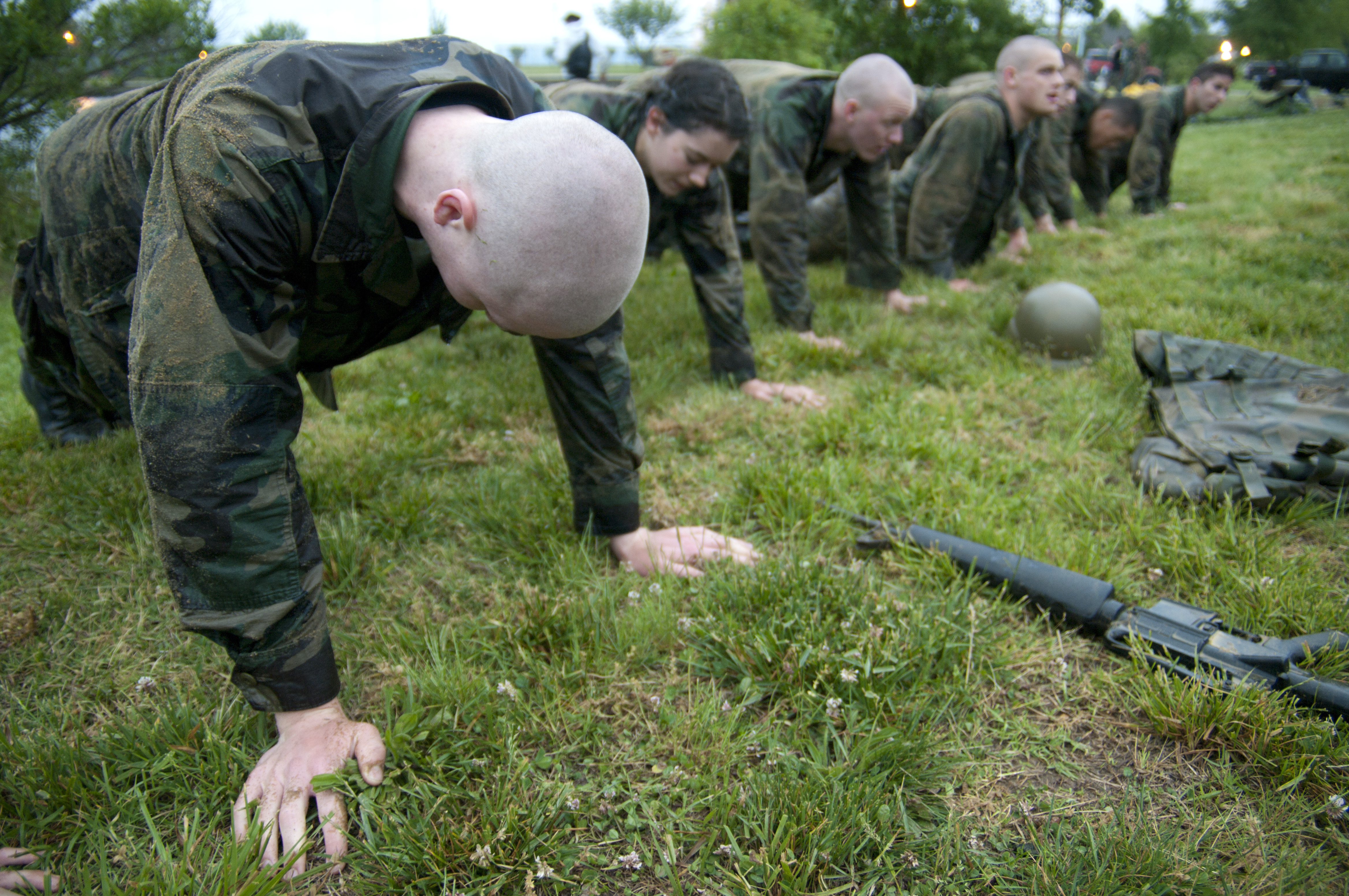 Eine Gruppe von Männern in Militäruniformen, die Liegestütze auf dem Gras machen, mit einer Waffe, einem Helm und anderen Gegenständen, die herumliegen, unter einem klaren blauen Himmel mit Bäumen, Lichtern und Fahrzeugen im Hintergrund.