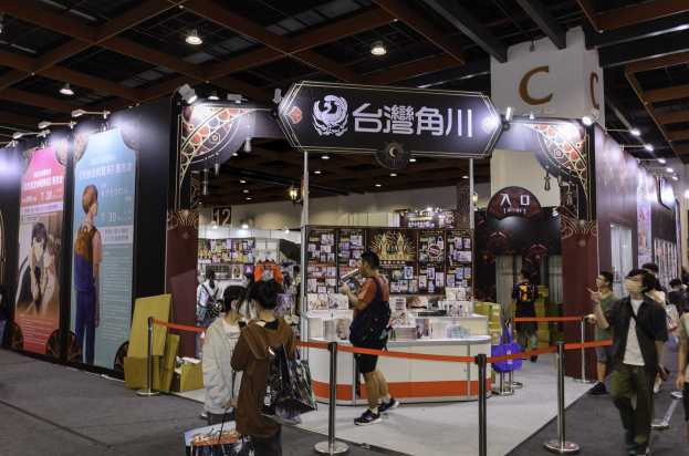 Menschen vor einem Messestand mit Werbematerialien, Taschen und Deckenbeleuchtung auf einer Veranstaltung, die wahrscheinlich mit der Tokyo Anime Expo 2019 in Verbindung steht.
