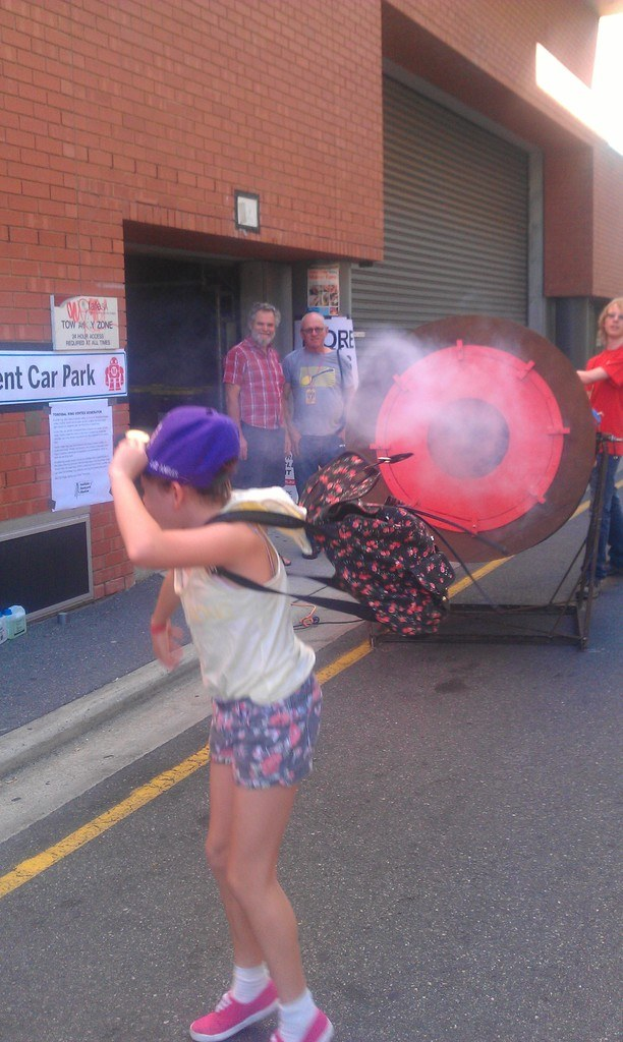 Ein Kind mit einem Rucksack und einer Mütze steht auf einer Straße mit Rauch im Hintergrund, ein kreisförmiges Objekt auf einem Ständer, Menschen, Wände, Pfosten an der linken Wand und Gegenstände auf dem Gehweg.