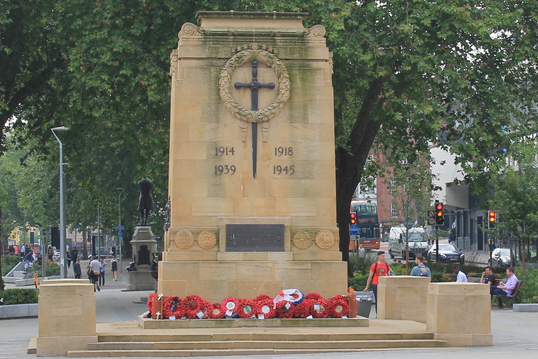 Kriegsdenkmal umgeben von Kränzen in einer Stadtstraße mit Menschen auf Bänken, Fahrzeugen, Verkehrsampeln, Laternenpfählen, Bäumen, Gebäuden und Himmel im Hintergrund.