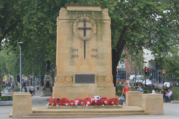 Kriegsdenkmal umgeben von Kränzen in einer Stadtstraße mit Menschen auf Bänken, Fahrzeugen, Verkehrsampeln, Laternenpfählen, Bäumen, Gebäuden und Himmel im Hintergrund.